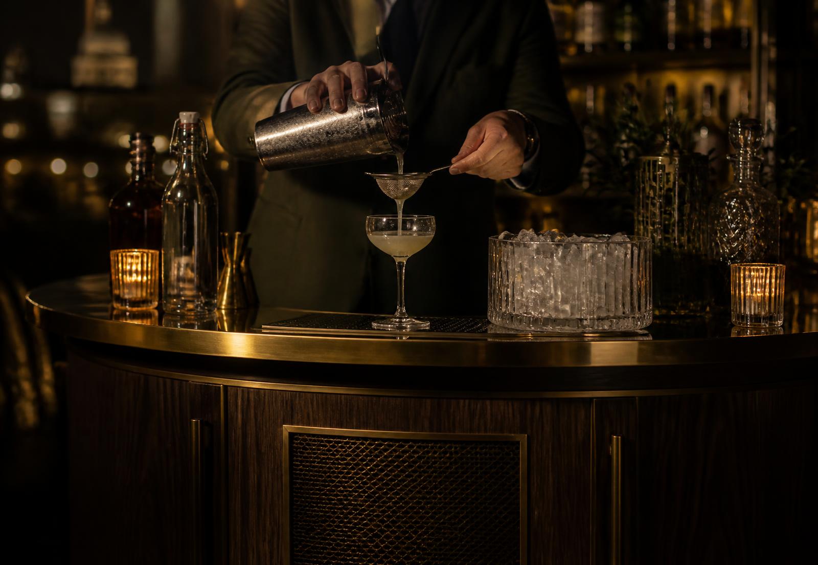 A bartender straining a cocktail into a coupe glass at a brass-topped London event bar